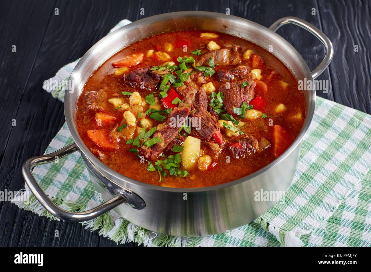 delicious hot hungarian goulash with beef meat, paprika, vegetables and csipetke - small egg pasta in stainless steel pot on black wooden table, class Stock Photo