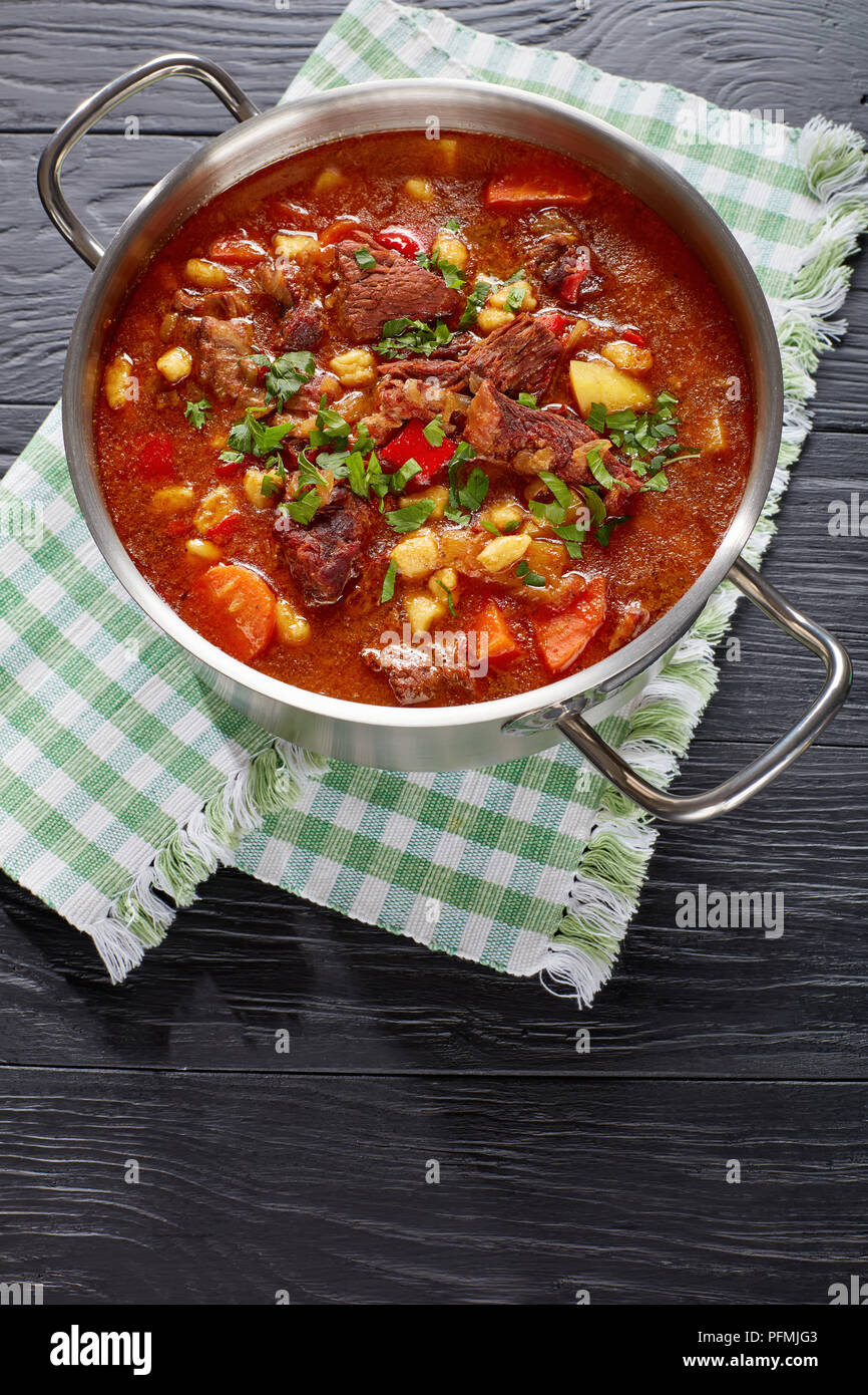 delicious hot hungarian goulash with beef meat, paprika, vegetables, csipetke, spices in stainless steel pot on black wooden table, authentic recipe,  Stock Photo