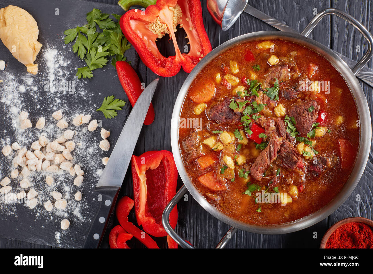 delicious hot beef hungarian goulash with paprika, small egg pasta, vegetables and spices in a pot. ingredients and kitchen ladle on table, classic re Stock Photo