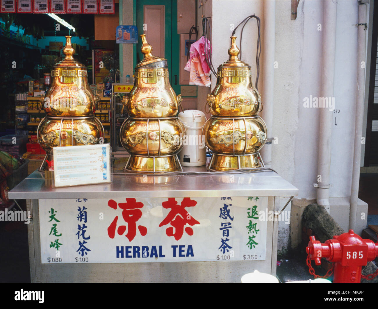 Singapore, Chinatown, Temple Street, decorative herbal tea dispensers, gold coloured metal decorated with Chinese script and dragons, lid loosely placed on central dispenser, standing on metal box, shop in background. Stock Photo