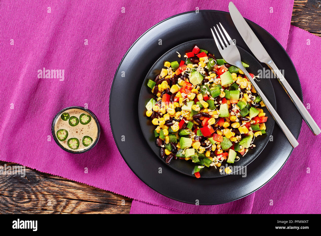 mexican style salad with avocado, black beans, canned corn, buckwheat sprouts, diced red and green pepper served on black plates with silver fork and  Stock Photo