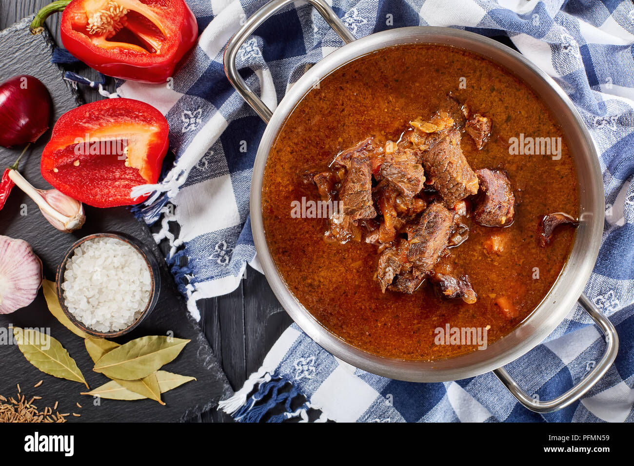 close-up of delicious homemade hot traditional Czech beef goulash in a stainless pan with kitchen towel on black wooden table with ingredients on cutt Stock Photo