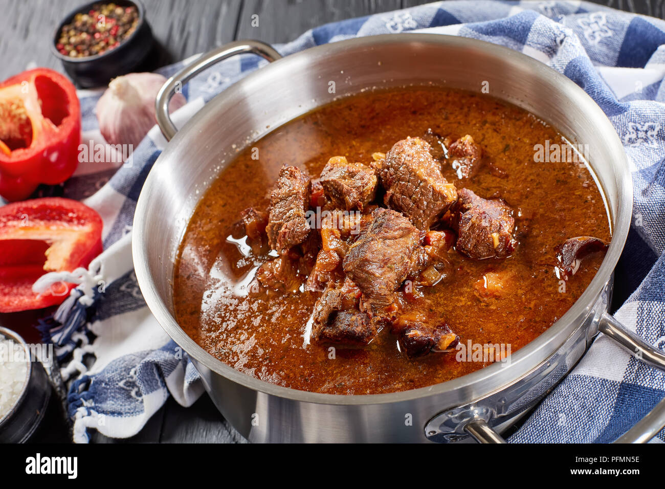 delicious homemade hot traditional Czech beef goulash in a stainless pan with kitchen towel on black wooden table with ingredients on cutting board, v Stock Photo