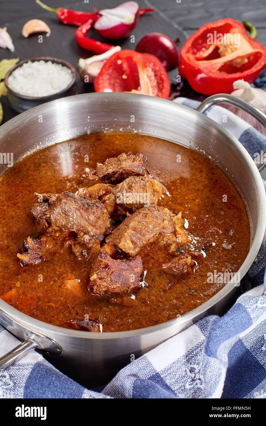 delicious homemade hot traditional Czech beef goulash in a stainless pan with kitchen towel on black wooden table with ingredients at background, vert Stock Photo