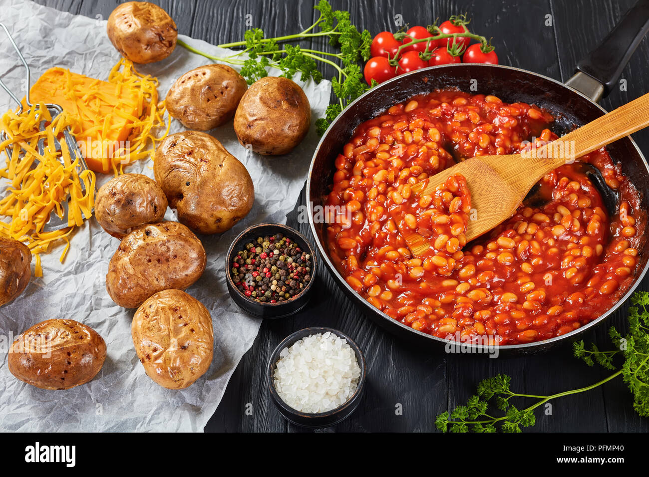 bean with tomato sauce in a skillet. baked potato or jacket potatoes with golden brown crispy skin on a paper with cheddar cheese at background, horiz Stock Photo