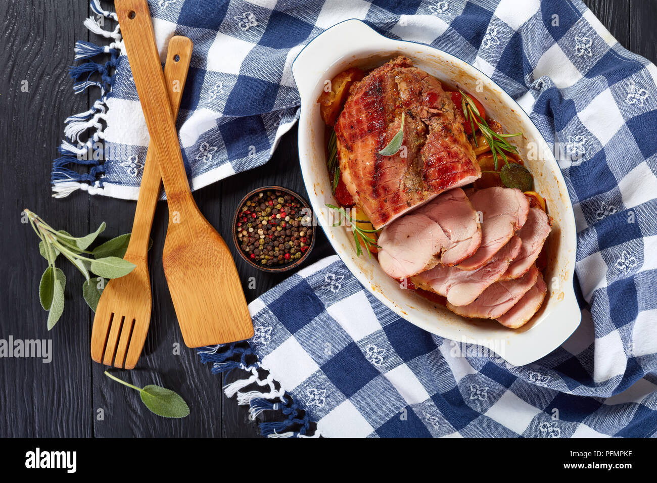 delicious roast pork cut in slices in a baking dish on wooden table with kitchen towel, fresh sage leaves and wooden spatulas, view from above Stock Photo