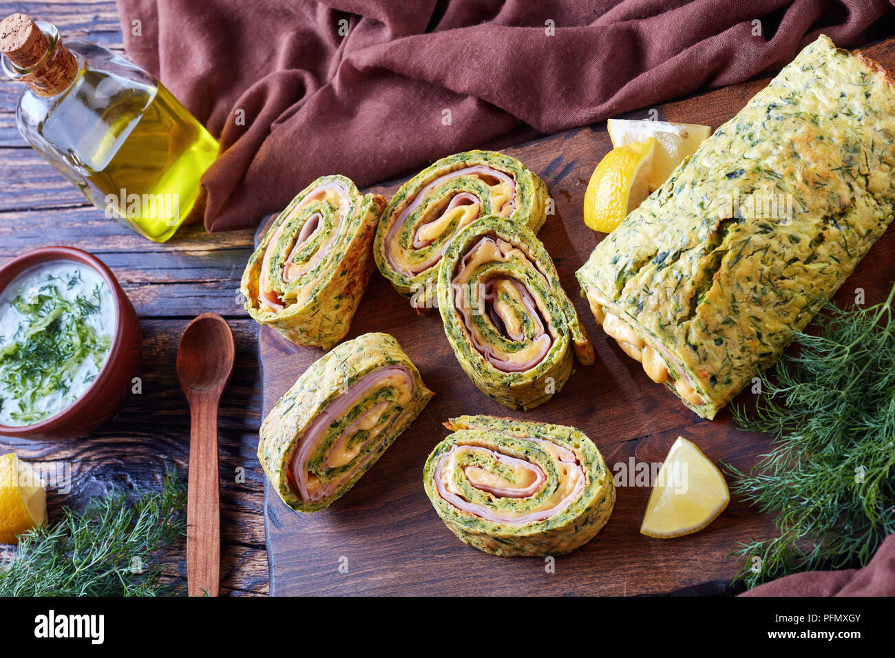 delicious zucchini roulade with melted cheese and boiled ham filling cut in slices on a cutting board with savory yogurt sauce in a bowl on a rustic t Stock Photo