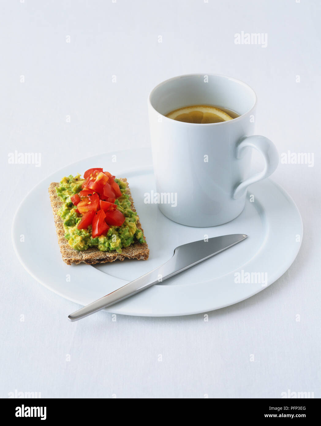 Mug of clear soup served on white ceramic plate, with rye crispbread topped with avocado spread and chopped tomatoes Stock Photo