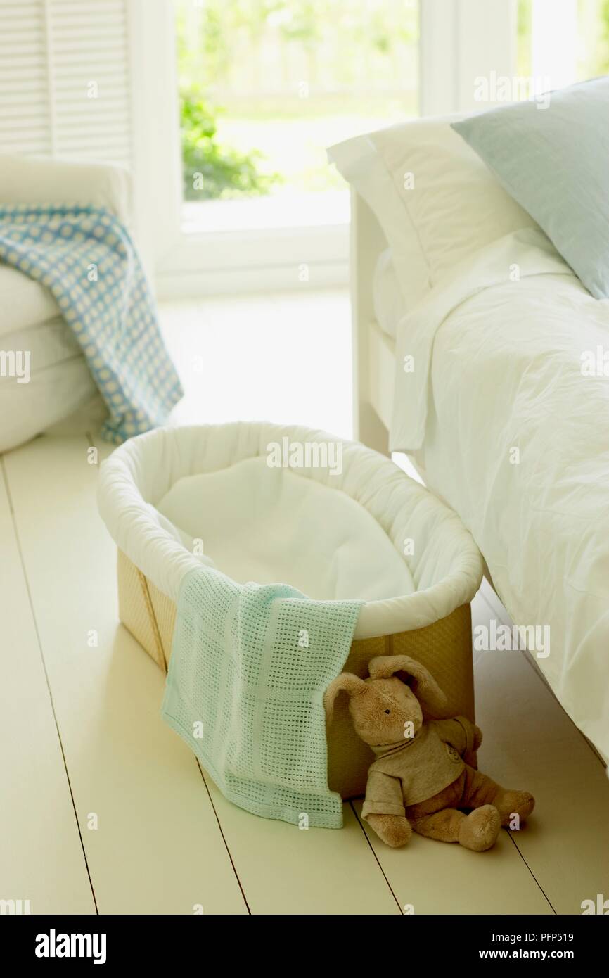 Moses basket, blankets, and soft toy rabbit next to bed, on wooden floor Stock Photo