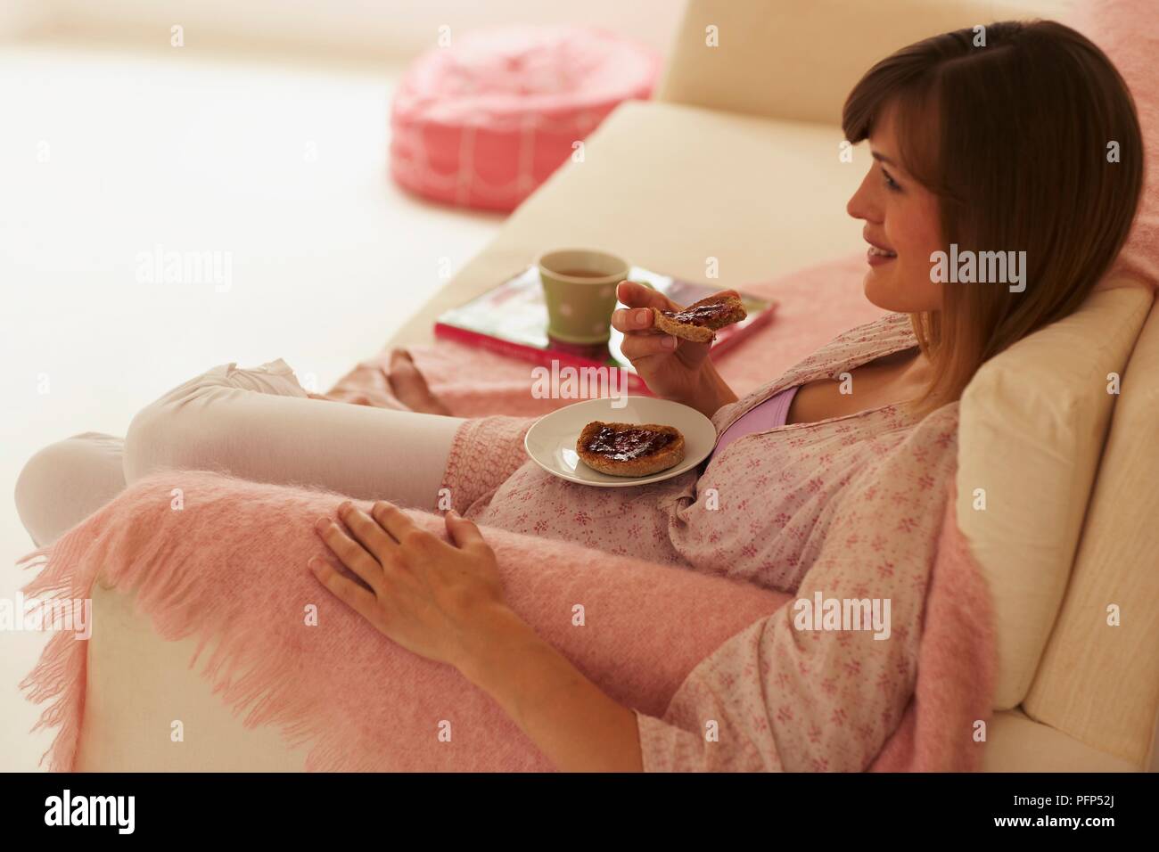 Pregnant woman sitting on sofa, plate balanced on her belly, eating slices of bread with jam, tea cup nearby, side view Stock Photo