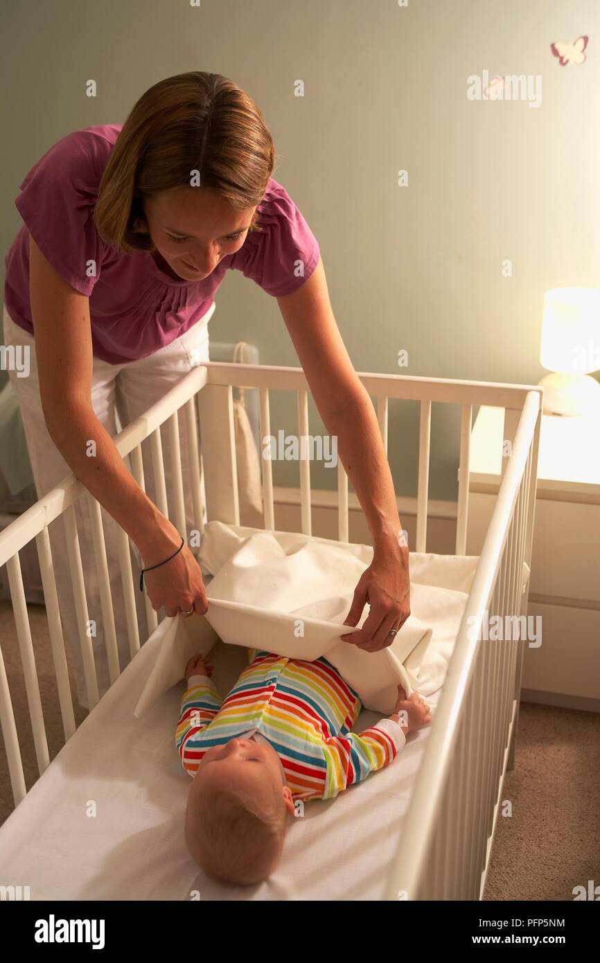 Baby boy in cot being covered with blanket by young woman Stock Photo