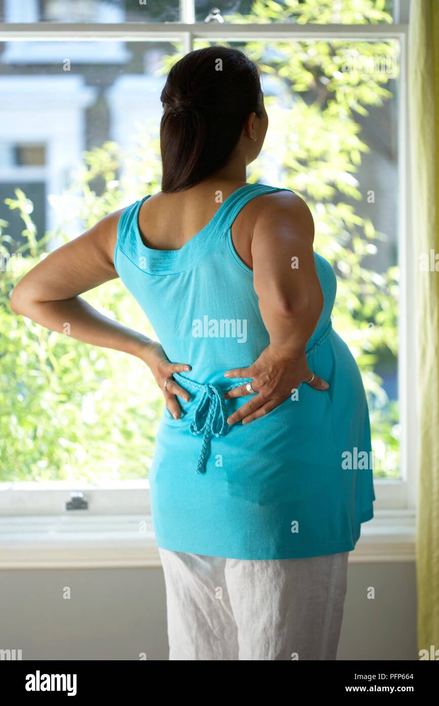 Pregnant woman standing by window, hands on her back, rear view Stock Photo