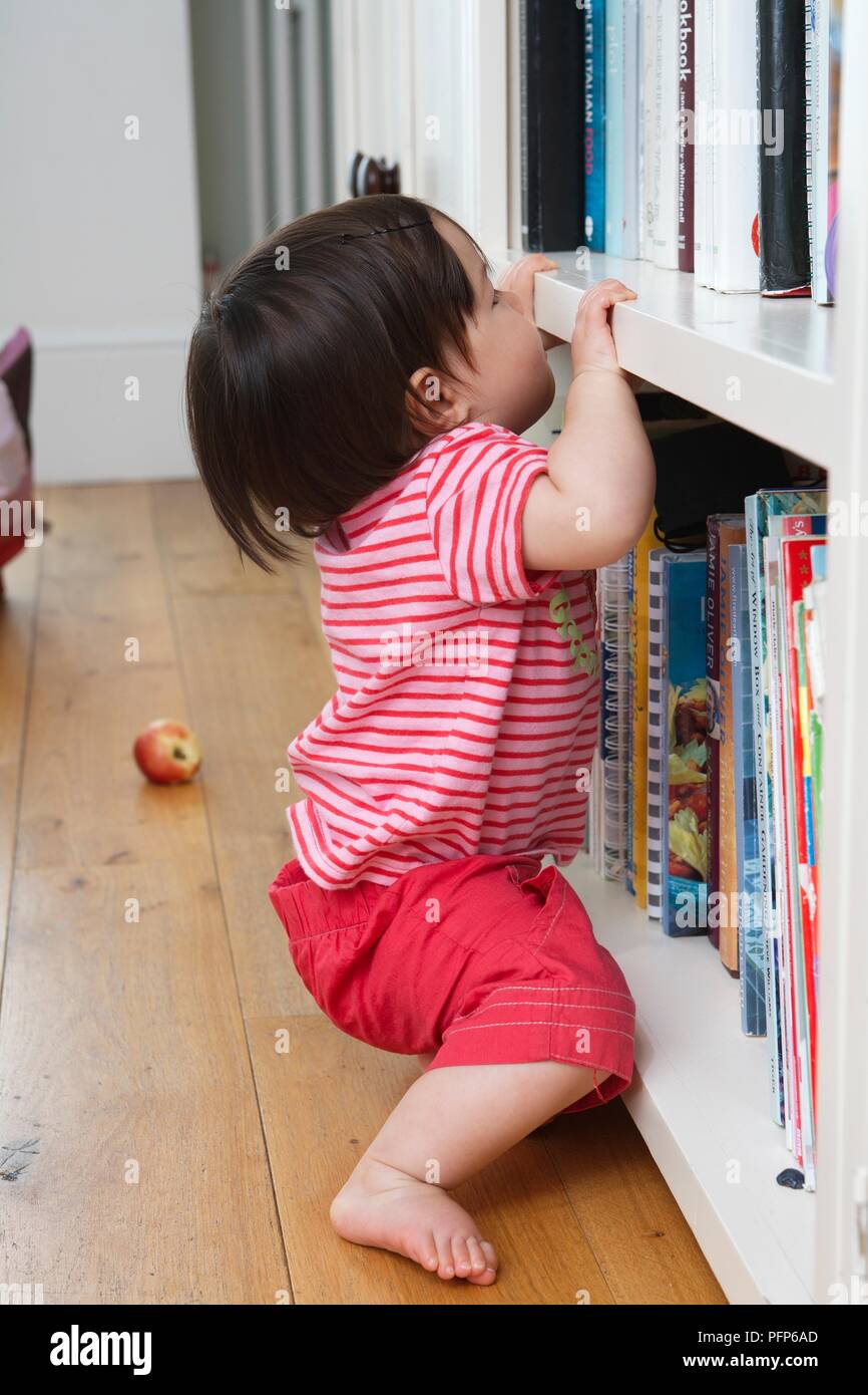 Baby Girl Pulling Herself Up On Bookshelf Side View Stock Photo