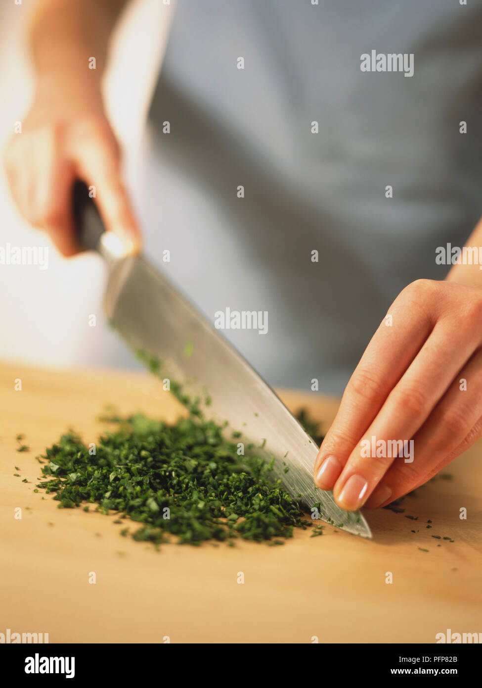 Petroselinum crispum, parsley, using large, sharp knife to cut parsley leaves, holding point of blade with flat fingers of non-cutting hand, chopping up and down in rocking motion on wooden work surface until herbs are finely cut. Stock Photo