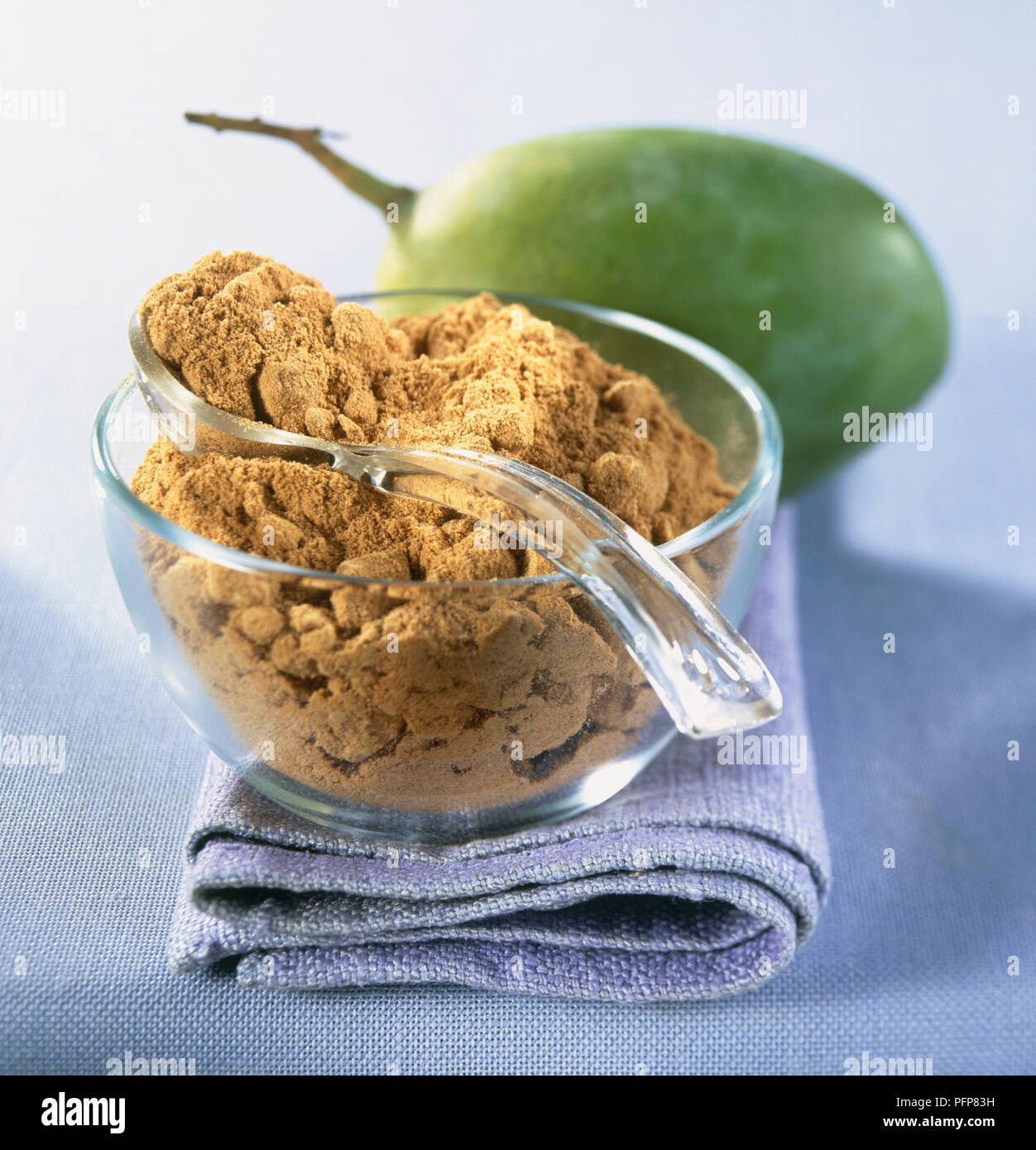 Powdered amchoor, a type of mango, on glass spoon and in glass bowl, with whole mango in the background Stock Photo