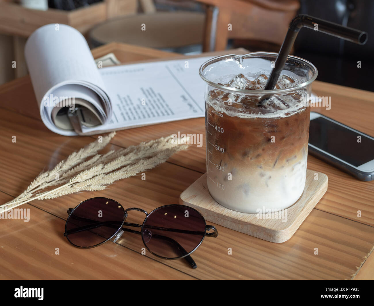 Ice coffee in beaker cup with black tube, mobile phone, sunglasses and paper on wooden table in vintage style cafe. coffee unique concept. Stock Photo