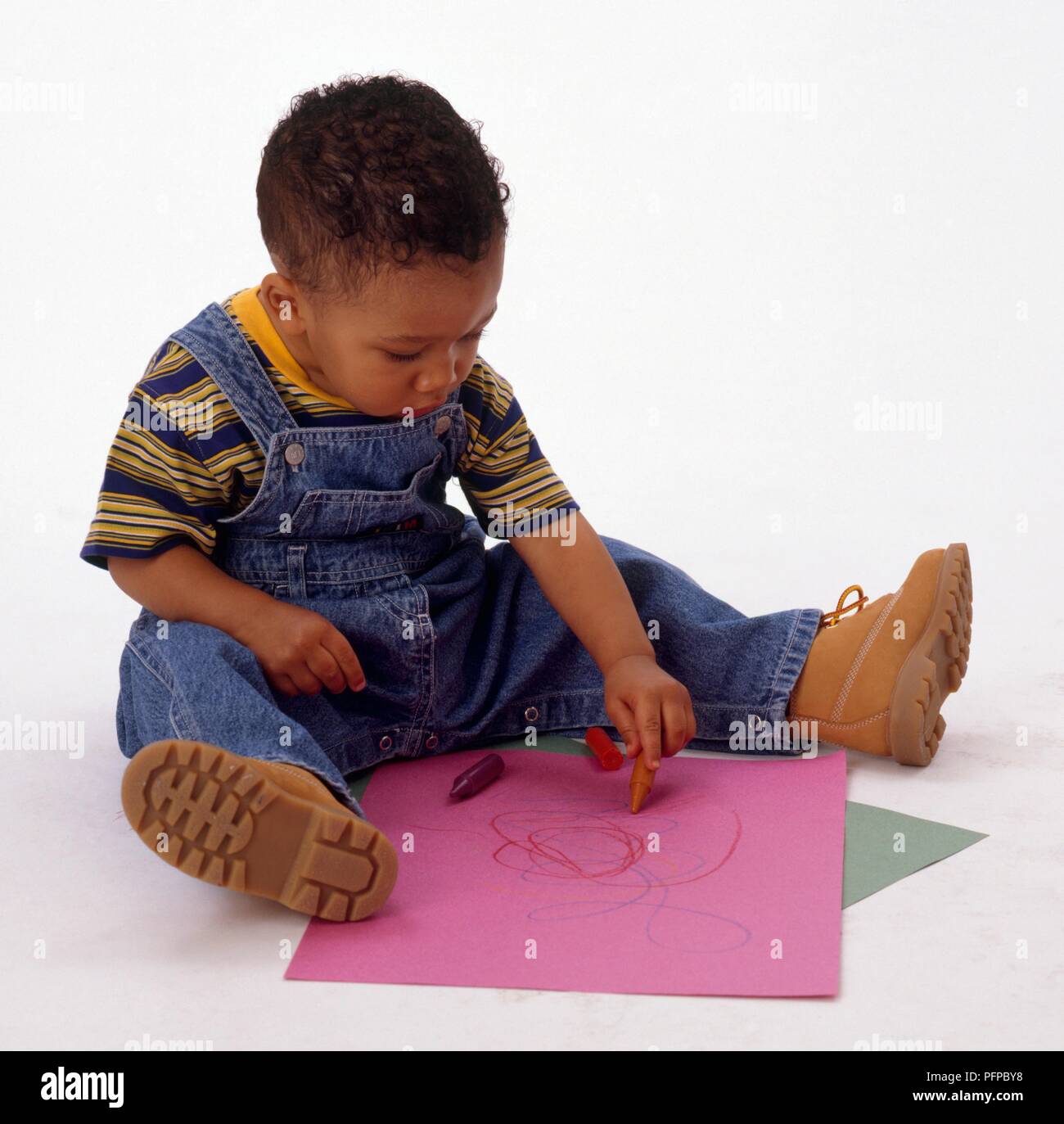 Boy stting in denim dungarees drawing on paper with crayons Stock Photo