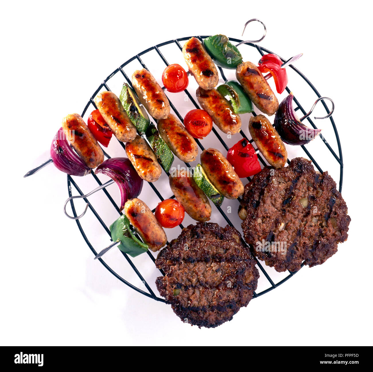 Two grilled beef burgers and sliced vegetables and cocktail sausages on skewers on wire wrack Stock Photo