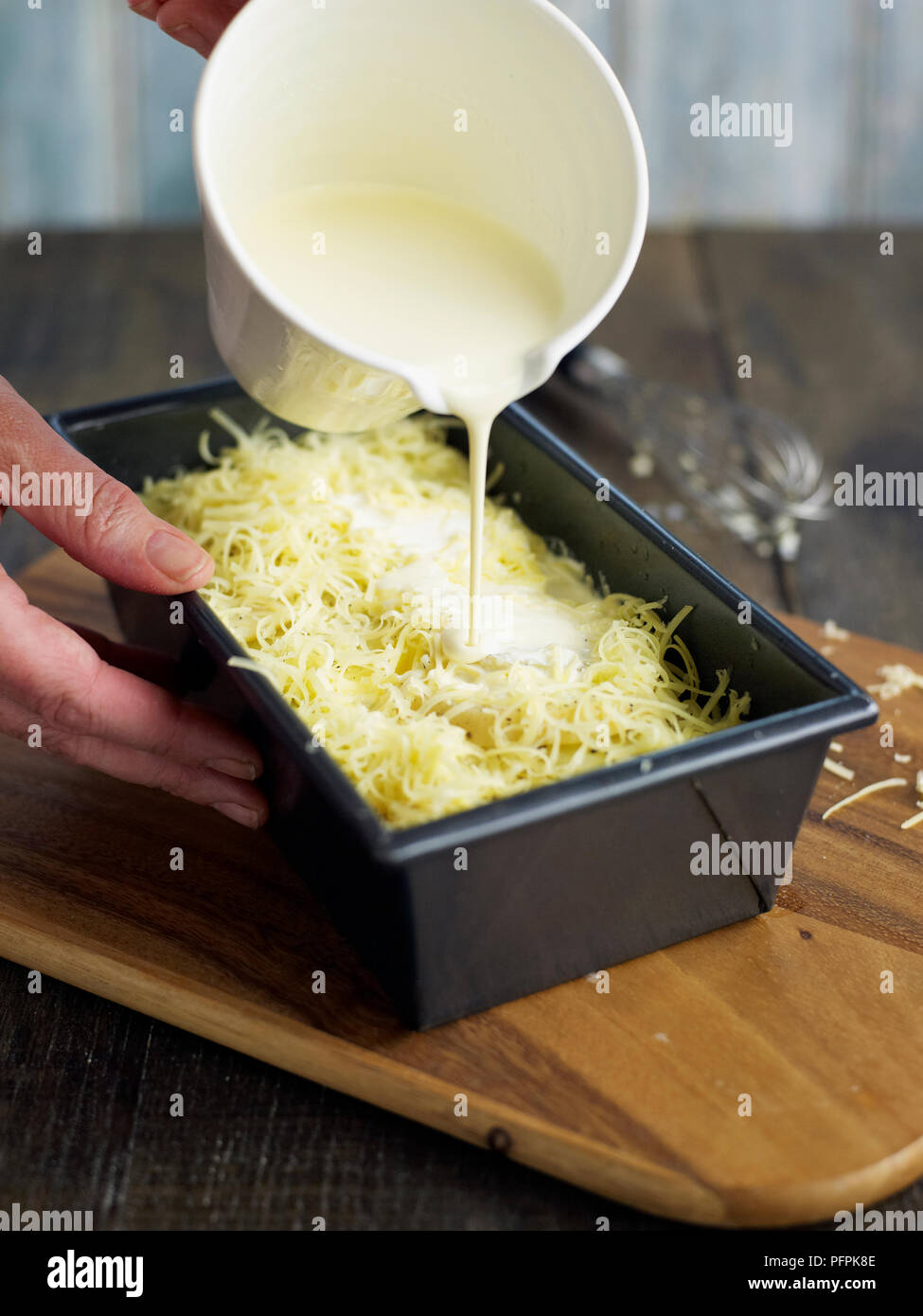 Pouring cream over grated cheese in loaf tin (making potato and celeriac gratin) Stock Photo