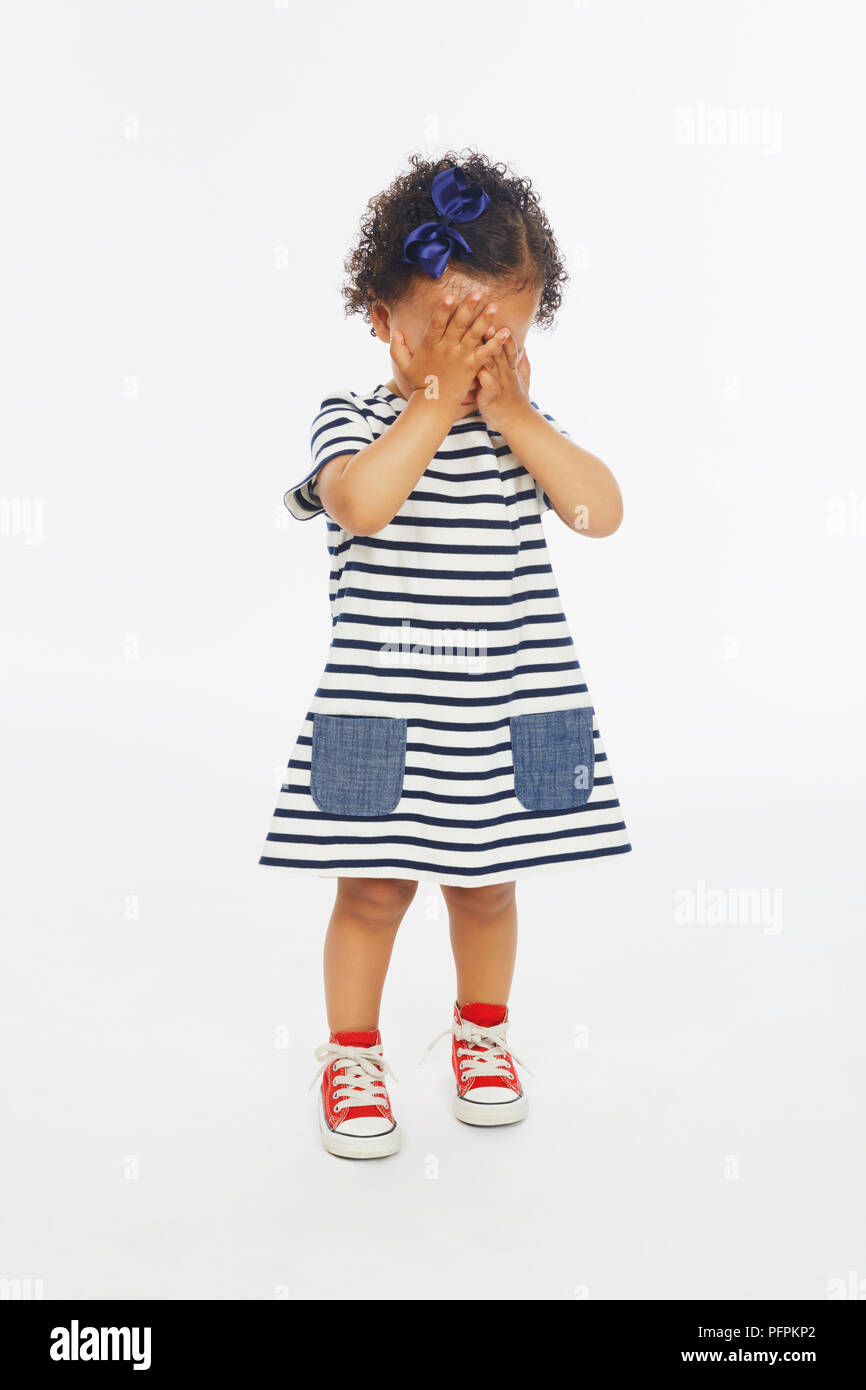 Little girl in stripey dress with both hands covering her face (Model age - 19 months) Stock Photo