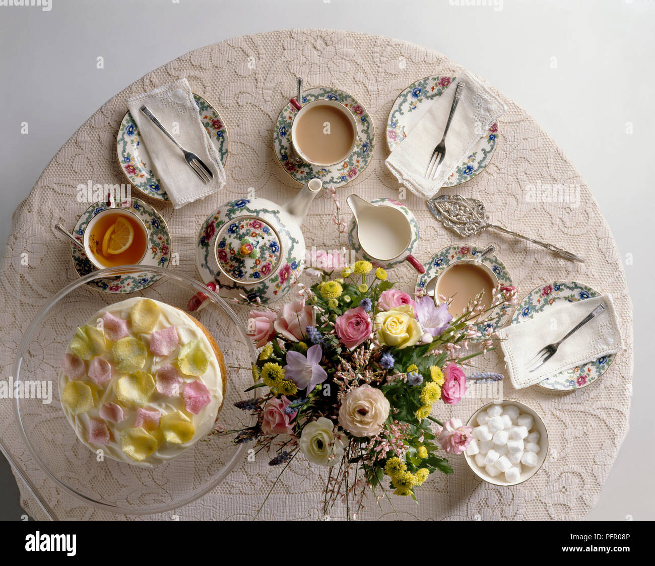 Table set for afternoon tea with bone china tea set, flower arrangement in centre, and cake decorated with rose petals Stock Photo
