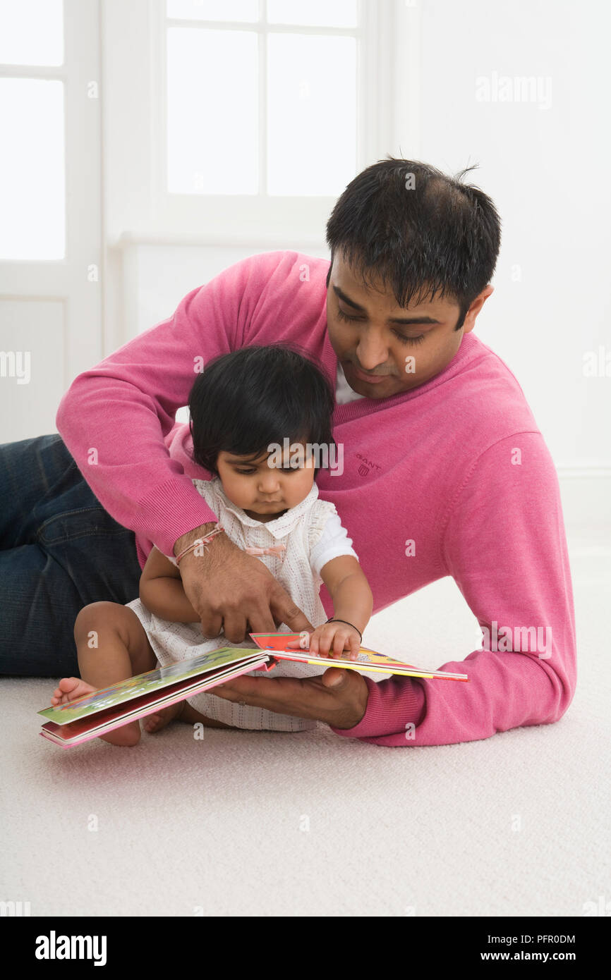 One year old baby girl sitting on floor with father as they both look at book Stock Photo