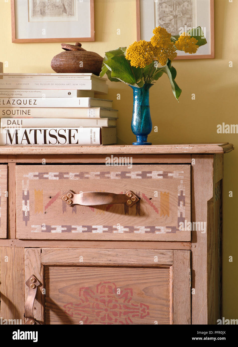 Wooden dresser with cupboard and drawers, stacked books on top, blue vase of yellow flowers, framed pictures on yellow wall behind. Stock Photo