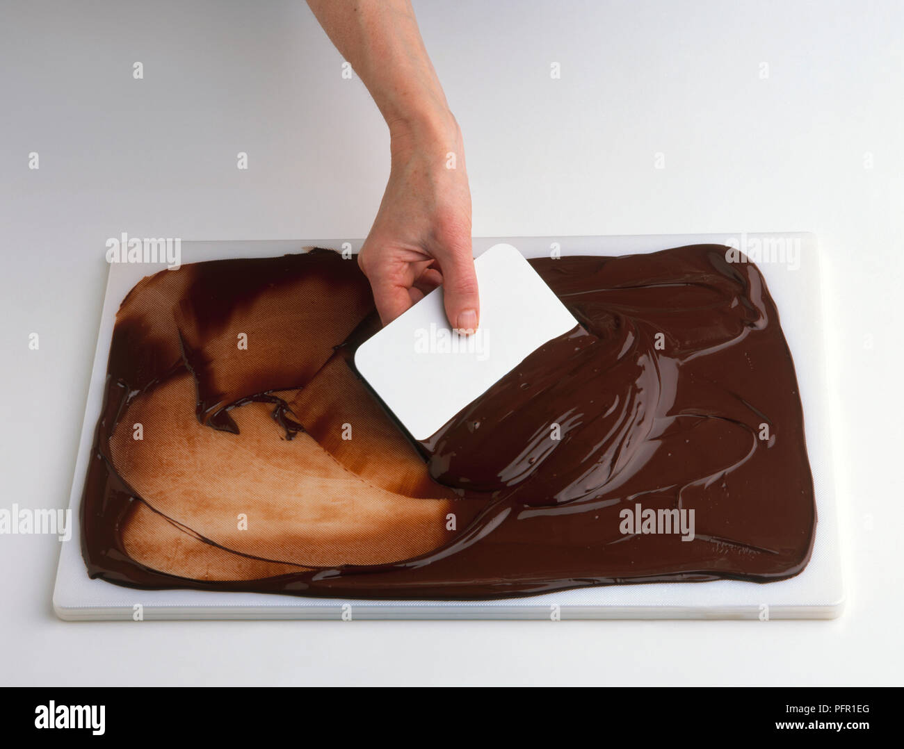 Man using plastic scraper to spread melted dark chocolate on chopping board, close-up Stock Photo