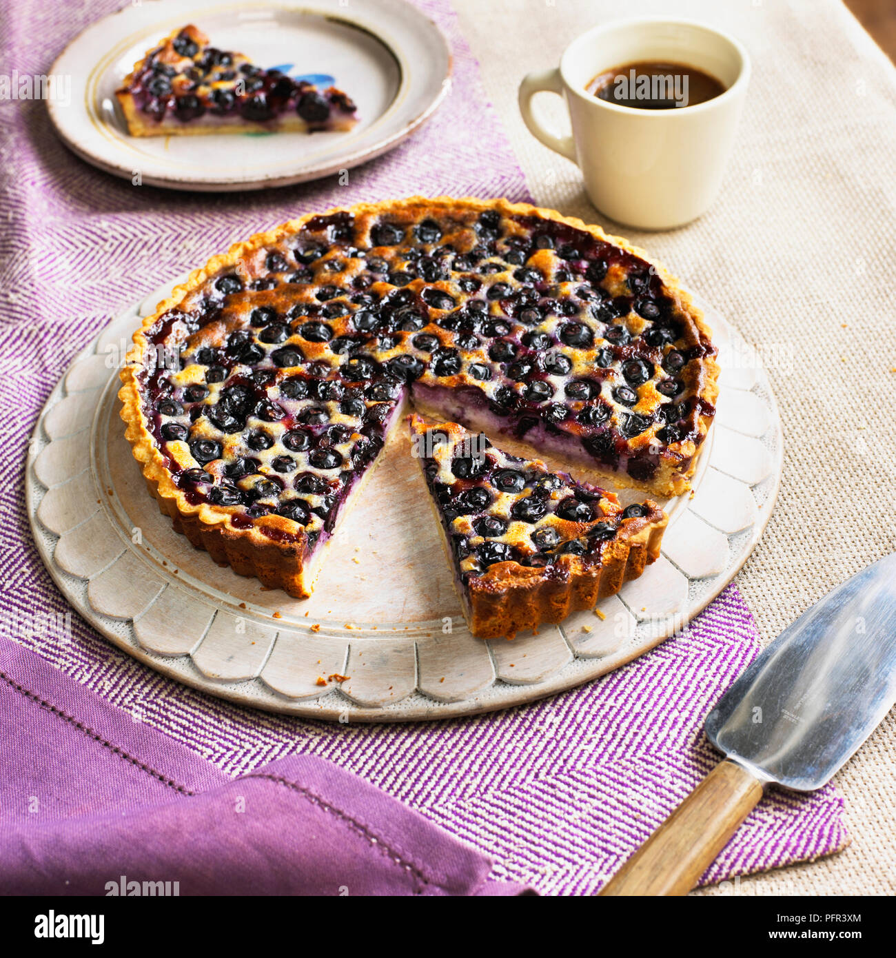 Blueberry cream cheese tart with slice cut away, cake server, coffee cup and separate slice on plate nearby Stock Photo