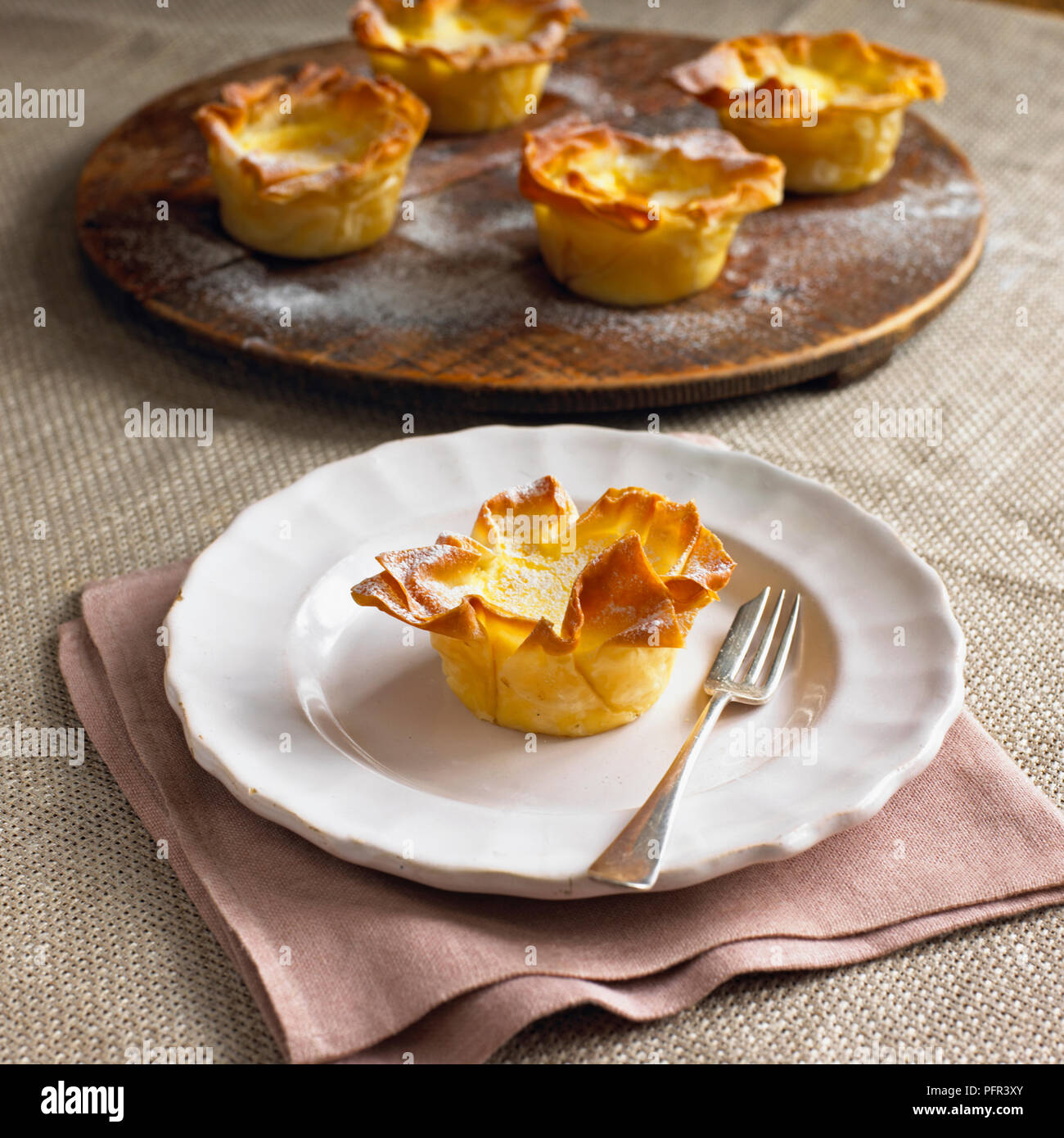 Cardamom custard filo tartlet on plate, with dessert fork, more tartlets on wooden board in the background Stock Photo