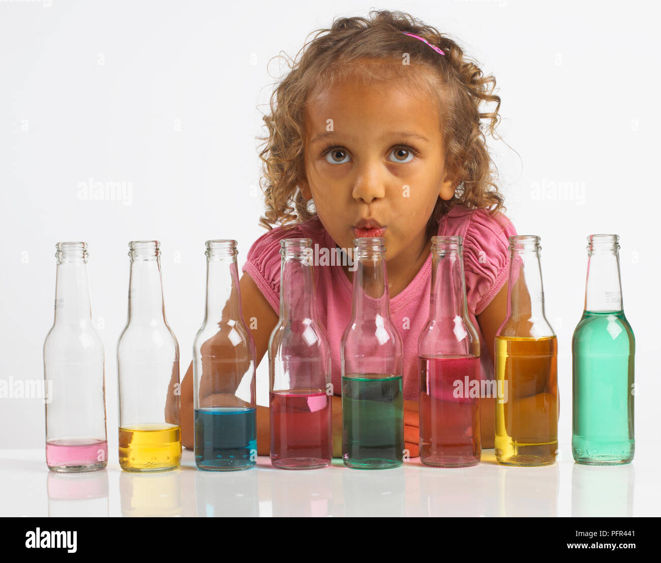 Girl playing 'bottle pipes', blowing into bottle which is part of a row of bottles filled with different amounts of coloured water, 4 years Stock Photo