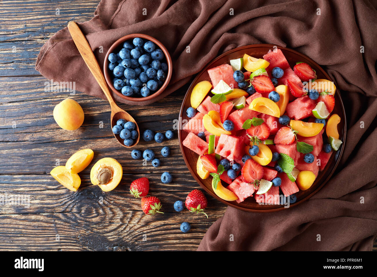 delicious colorful fresh fruit salad with watermelon, blueberries, peach slices, strawberry and lime on a clay plate on an old rustic table with ingre Stock Photo