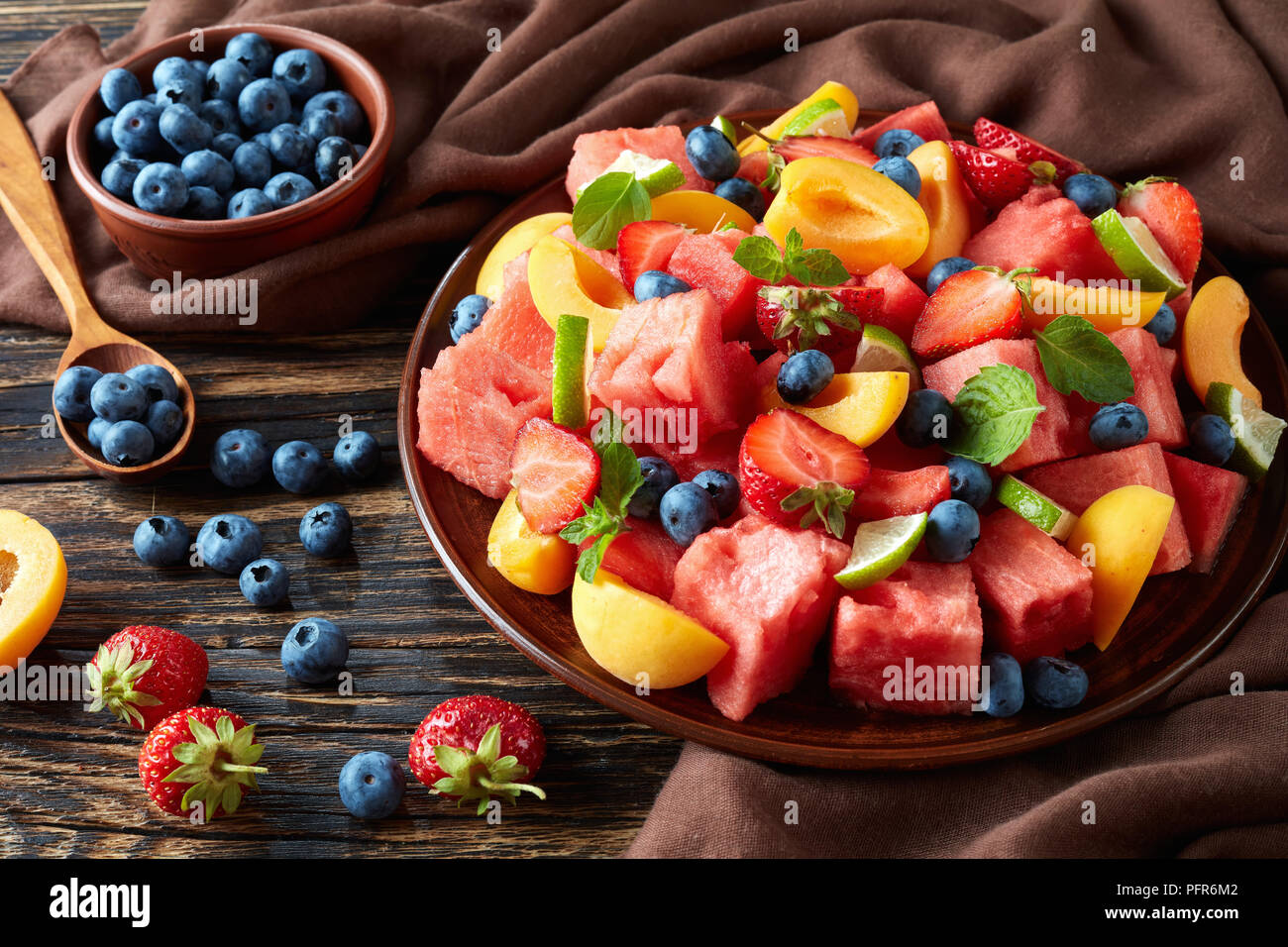 delicious colorful fresh fruit salad with watermelon, blueberries, peach slices, strawberry and lime on a clay plate on an old rustic table with ingre Stock Photo