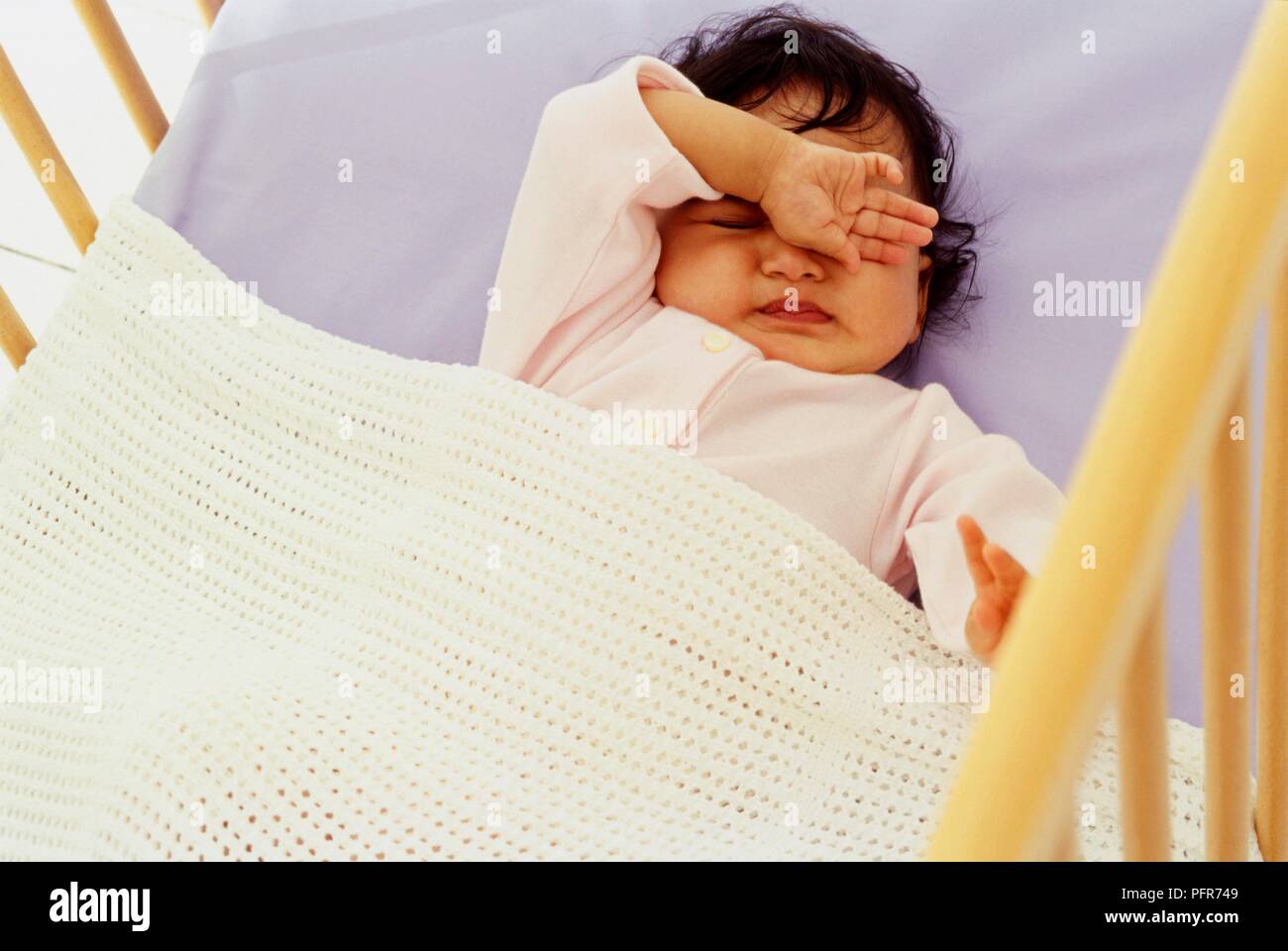 Baby lying on back in cot with one arm raised and rubbing her eyes