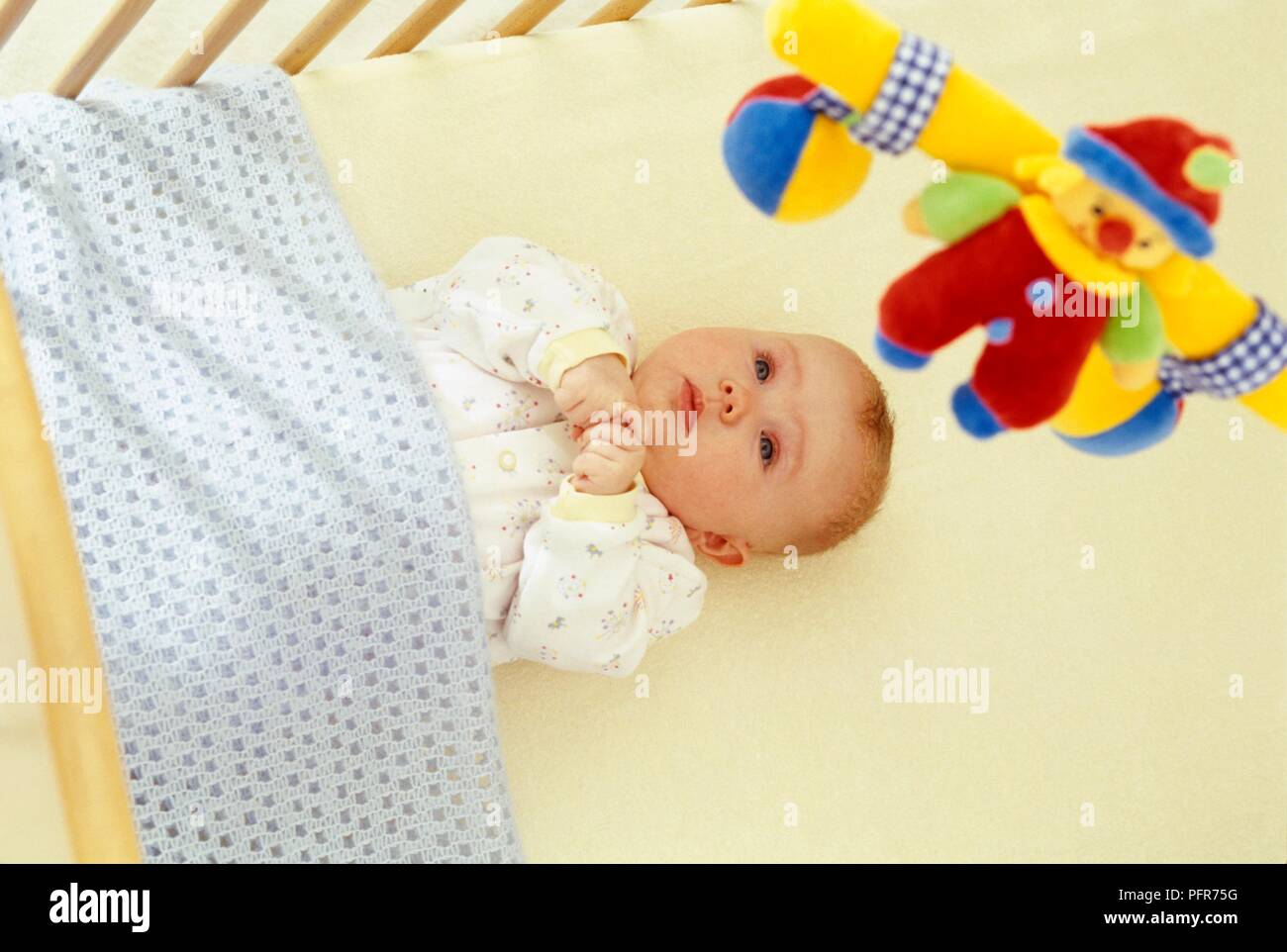 Baby lying in cot covered in blanket looking up to colourful clown mobile Stock Photo