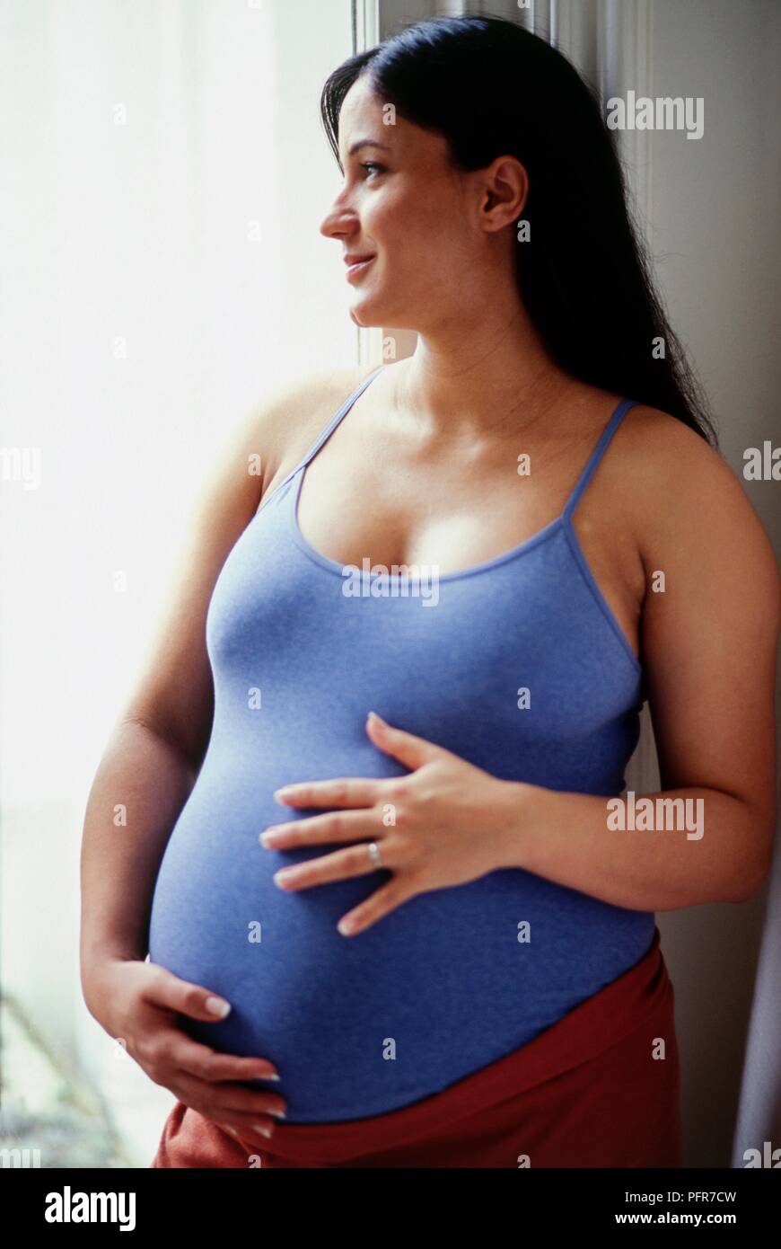 Pregnant woman standing next to window holding her belly Stock Photo