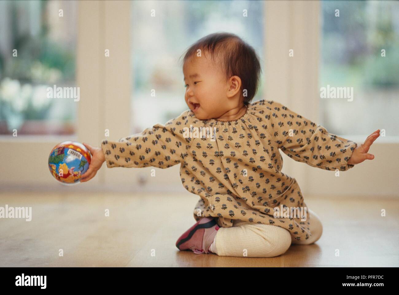 Baby girl kneeling on the floor and extending ball outwards with one