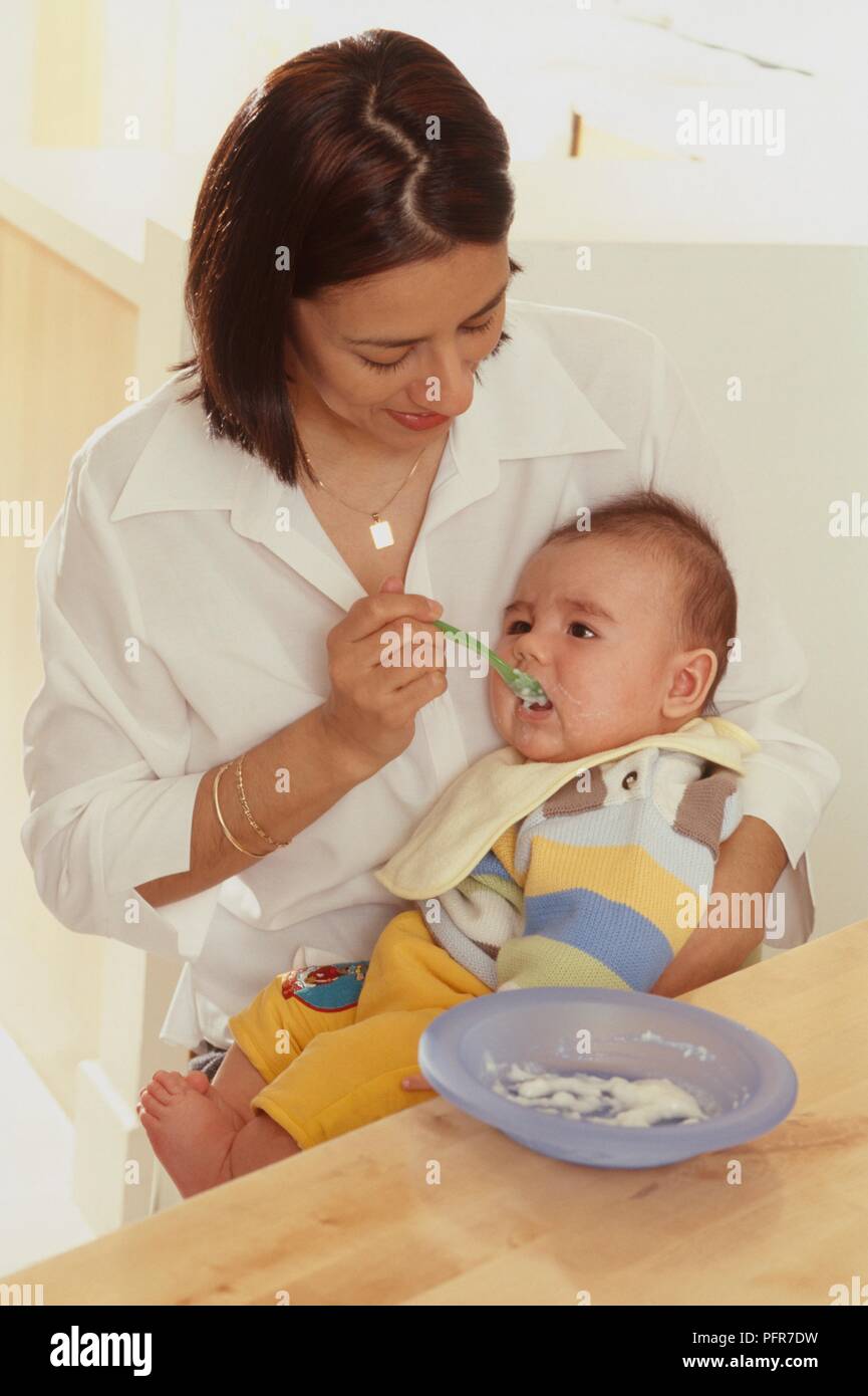 Woman Seated At Table With Baby Boy On Her Lap Feeding Him With Spoon woman-seated-at-table-with-baby-boy-on-her-lap-feeding-him-with-spoon