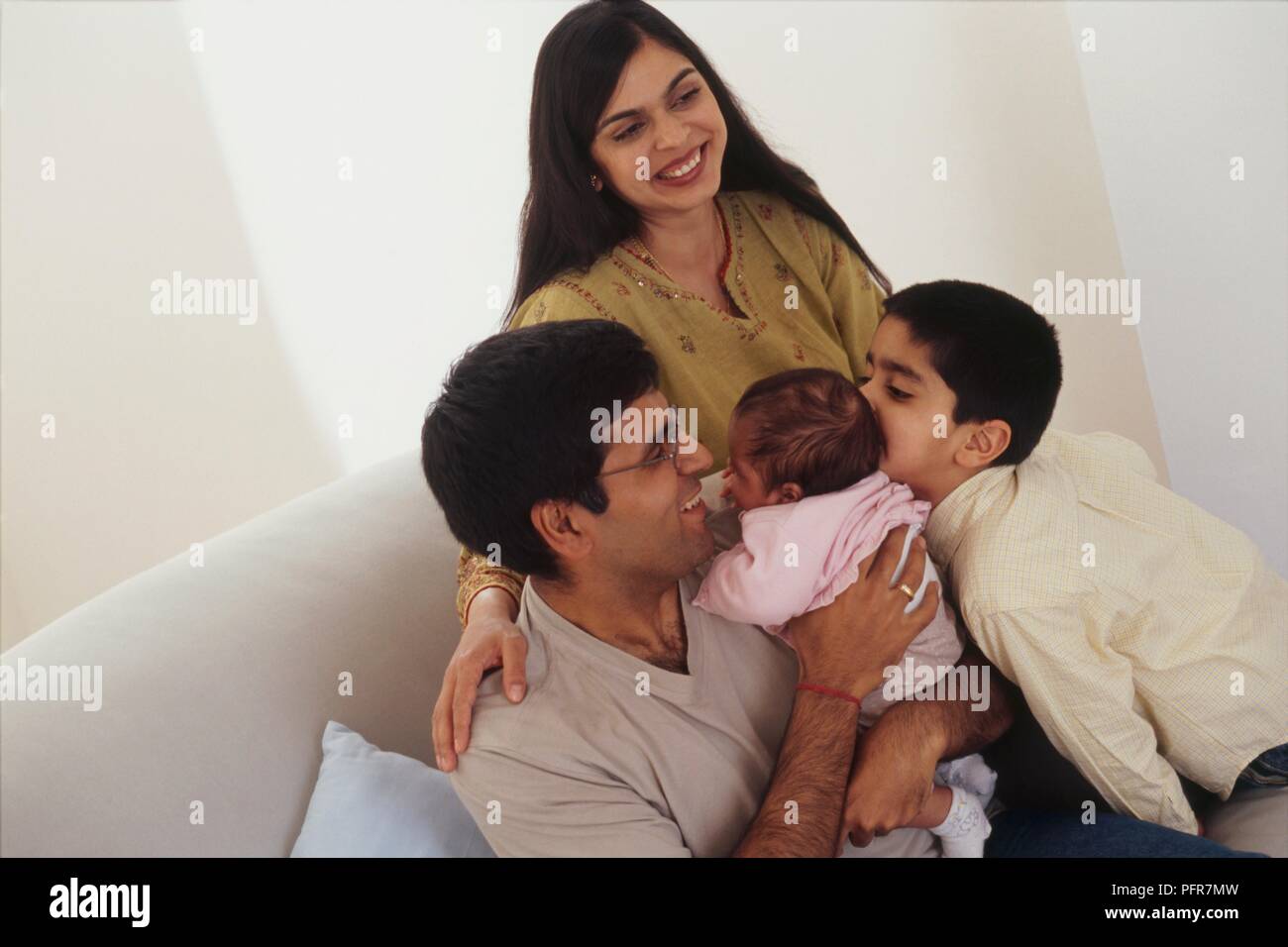 A man, woman, boy and baby girl together on sofa, man holding the baby and boy kissing the baby on the side of the head Stock Photo