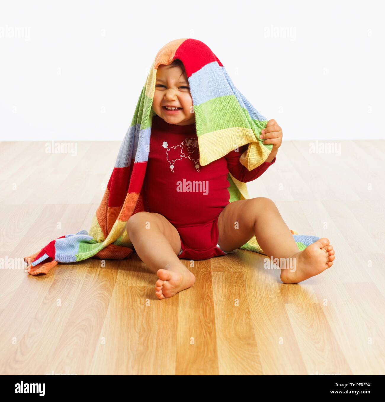 Baby girl sitting with a stripy blanket over her head, smiling Stock Photo