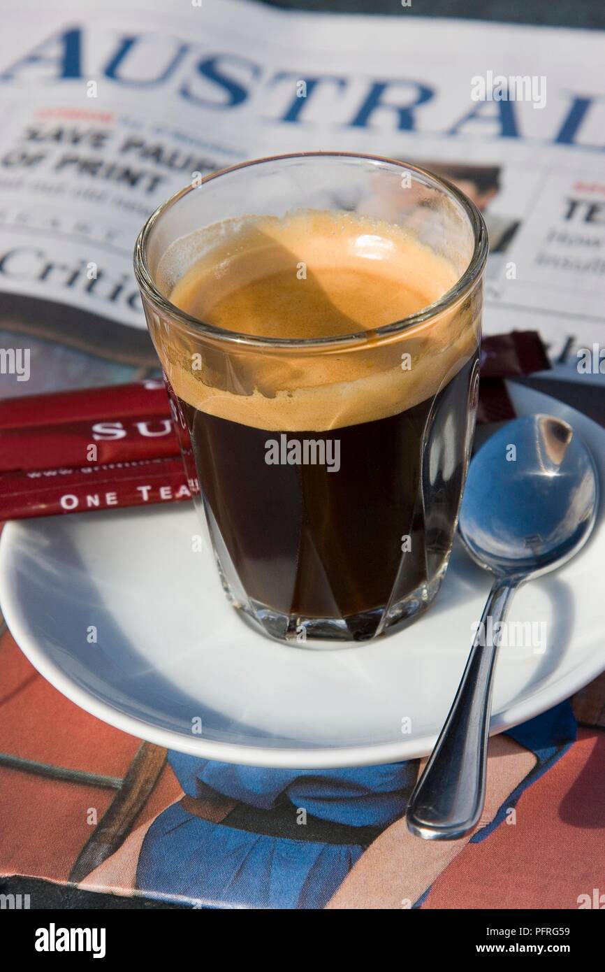 Australia, coffee in glass on saucer with spoon and sugar on table and newspaper in background Stock Photo