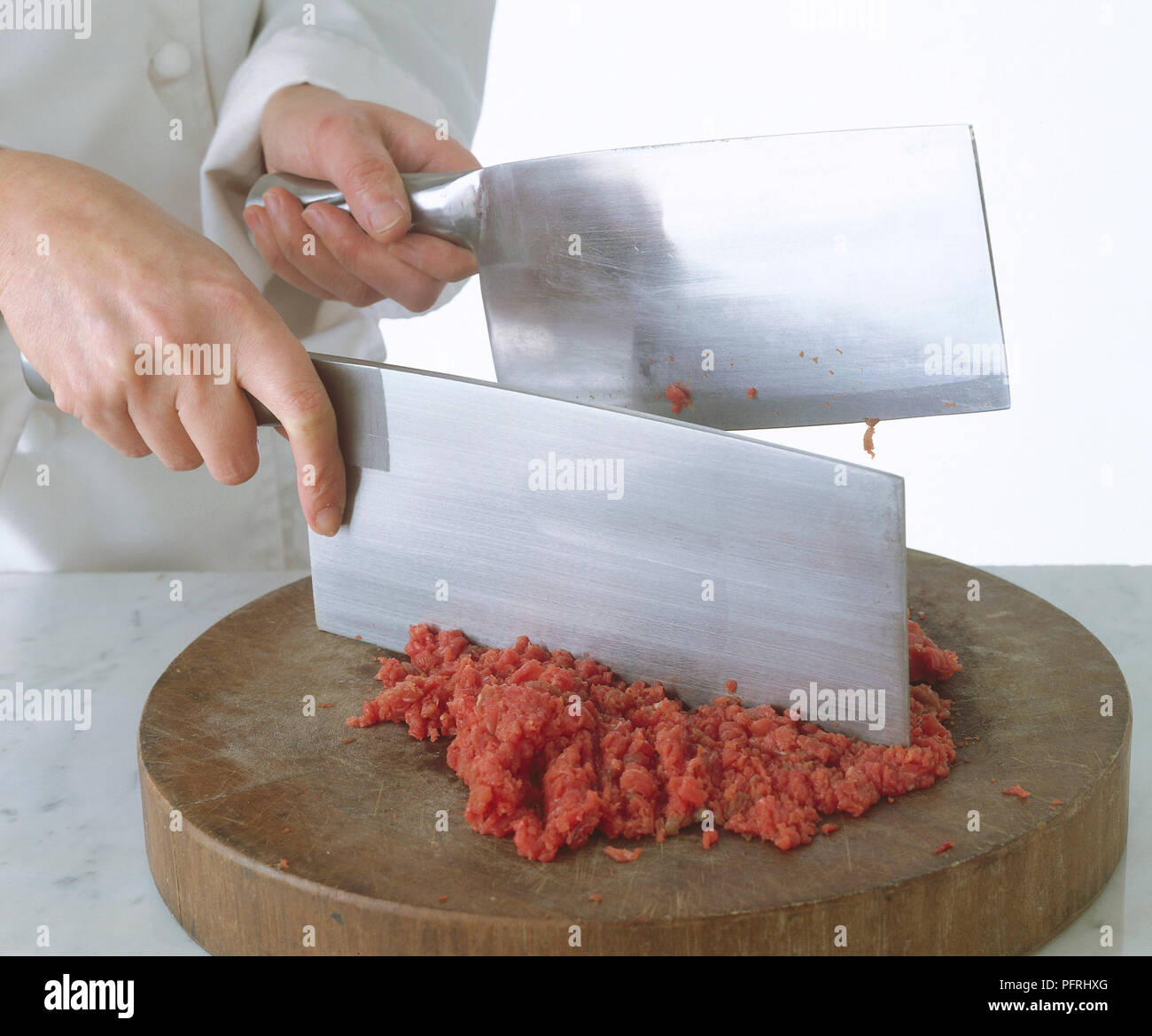 Chef using two meat cleavers to chop minced meat on wooden chopping board on kitchen worktop