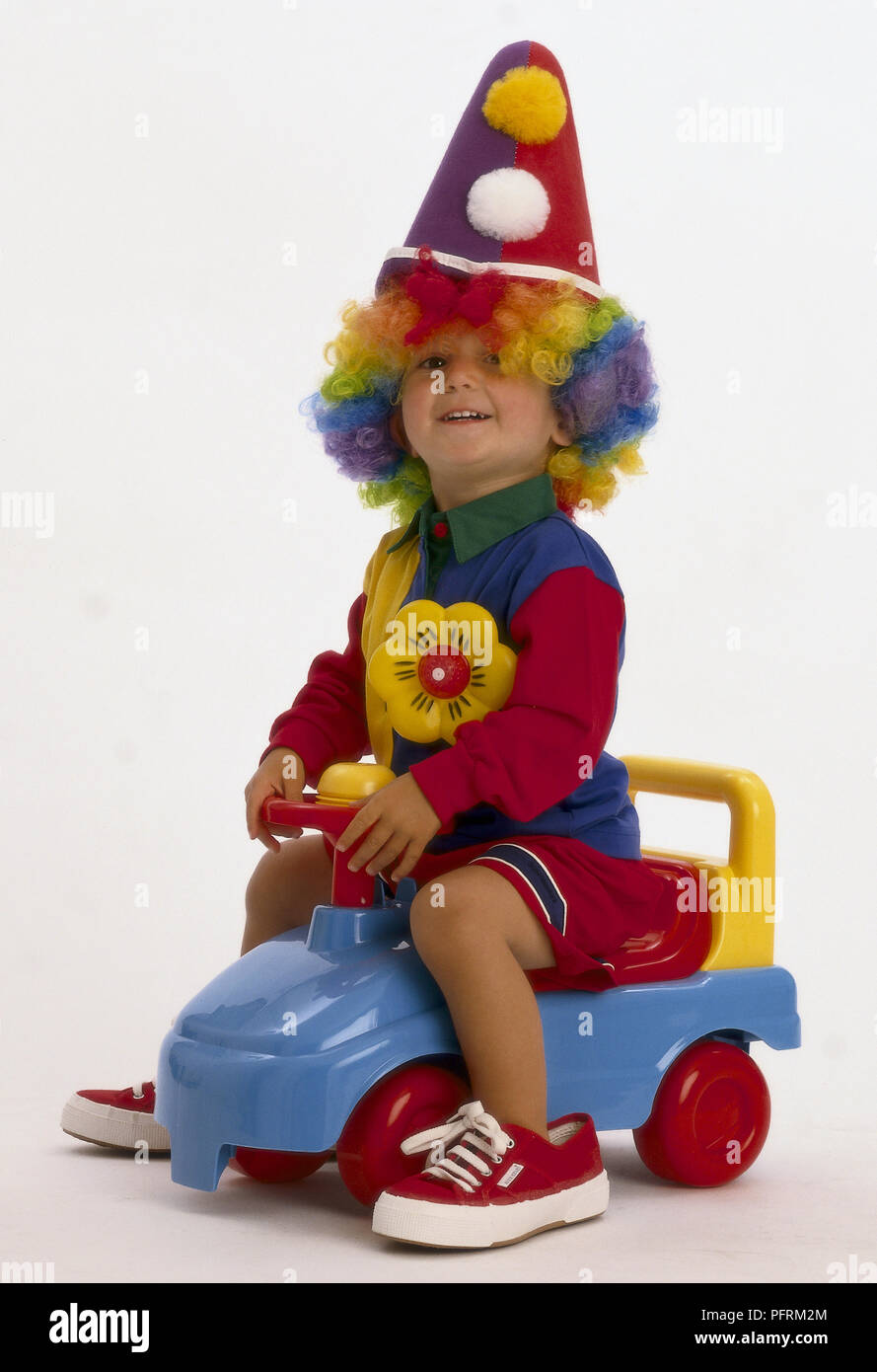 Smiling toddler wearing clown hat and multi coloured wig and oversized canvas shoes sitting on toy car Stock Photo