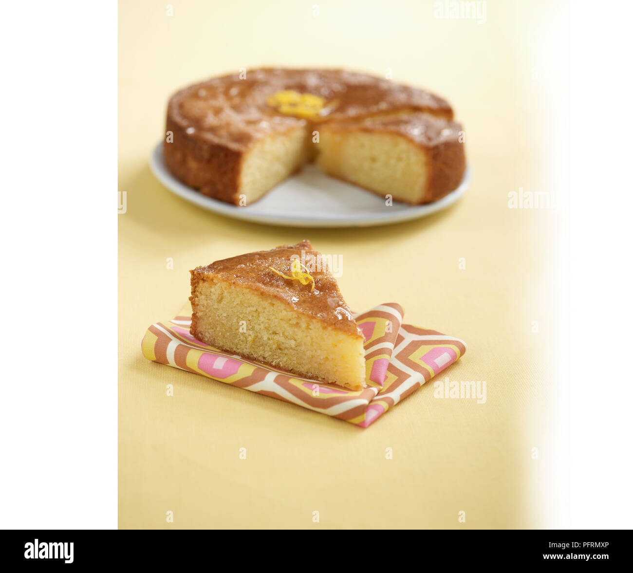 Lemon drizzle cake on plate and single slice on napkin in the foreground Stock Photo