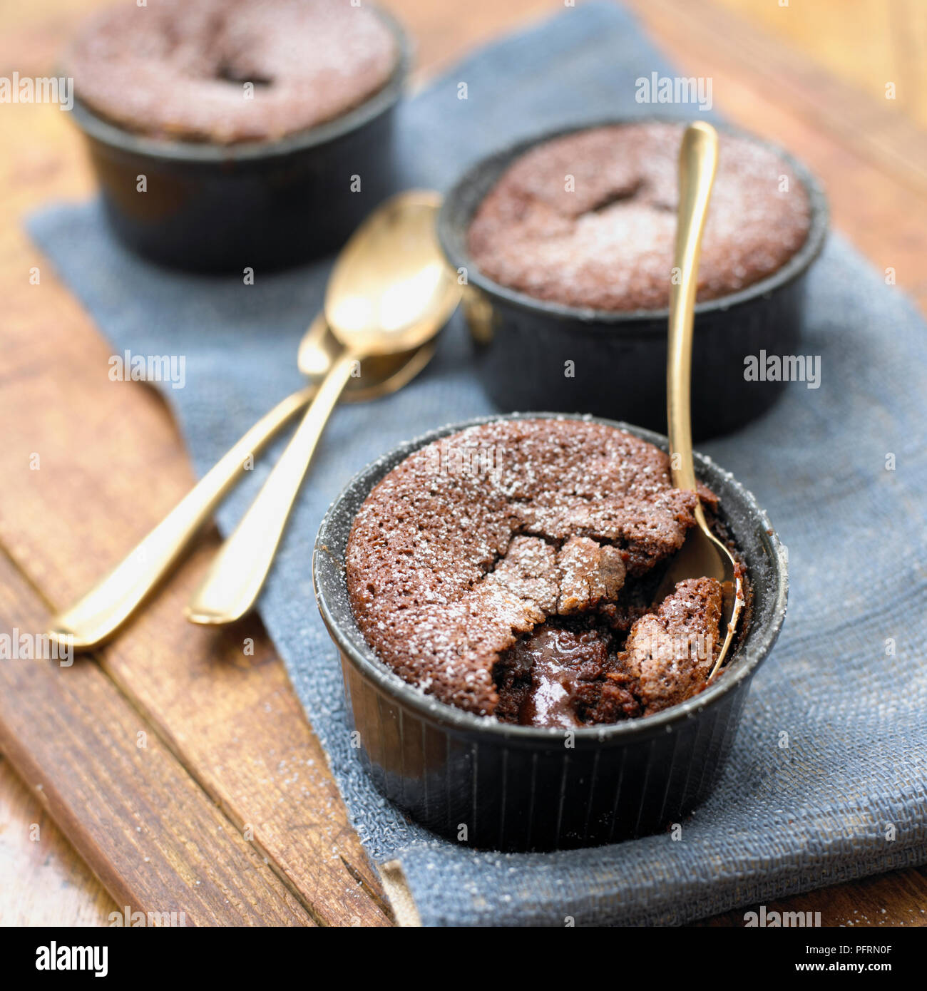 Hot dark chocolate fondant puddings in ramekins dusted with icing sugar, with spoons and one pudding partly eaten, close-up Stock Photo