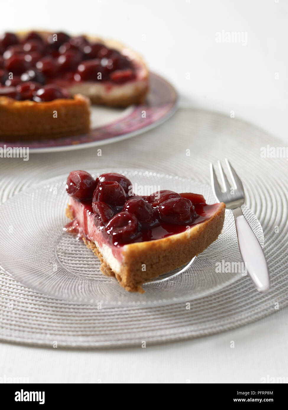 Cheery cheesecake slice on glass plate, with a dessert fork nearby Stock Photo