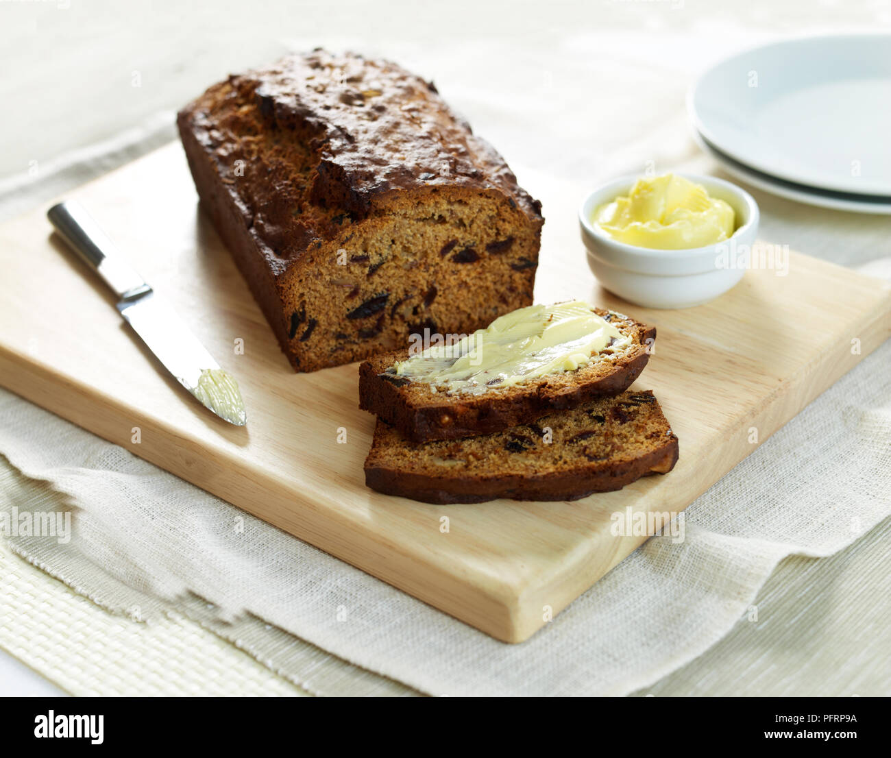 Date and walnut loaf with some slices cut away and one of the slices buttered, knife and butter nearby Stock Photo
