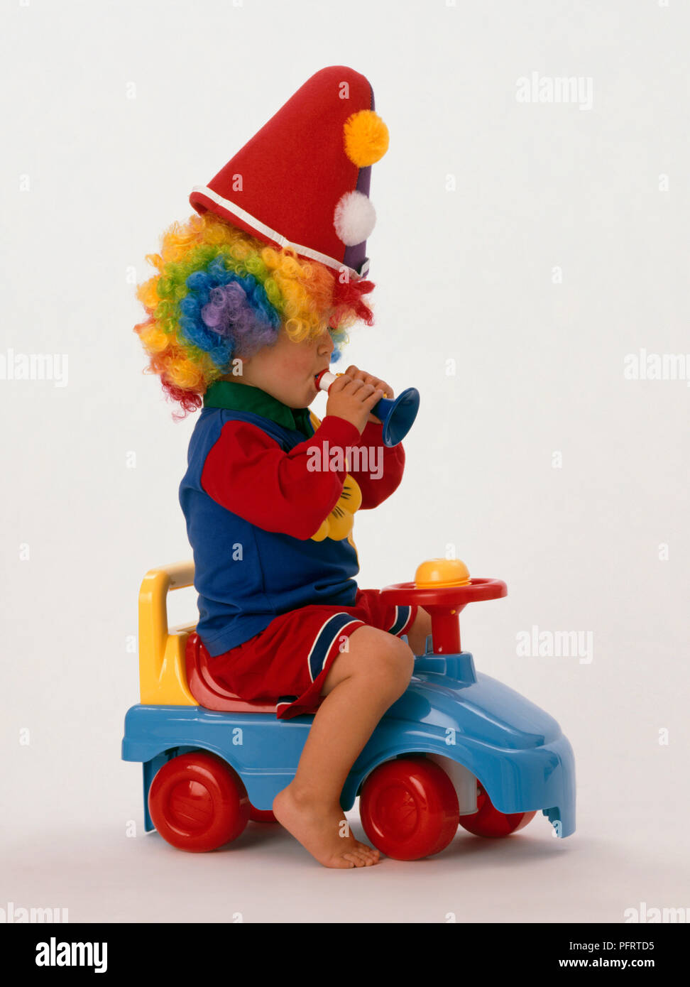 Toddler wearing clown hat and multi coloured wig sitting on toy car and blowing toy trumpet Stock Photo