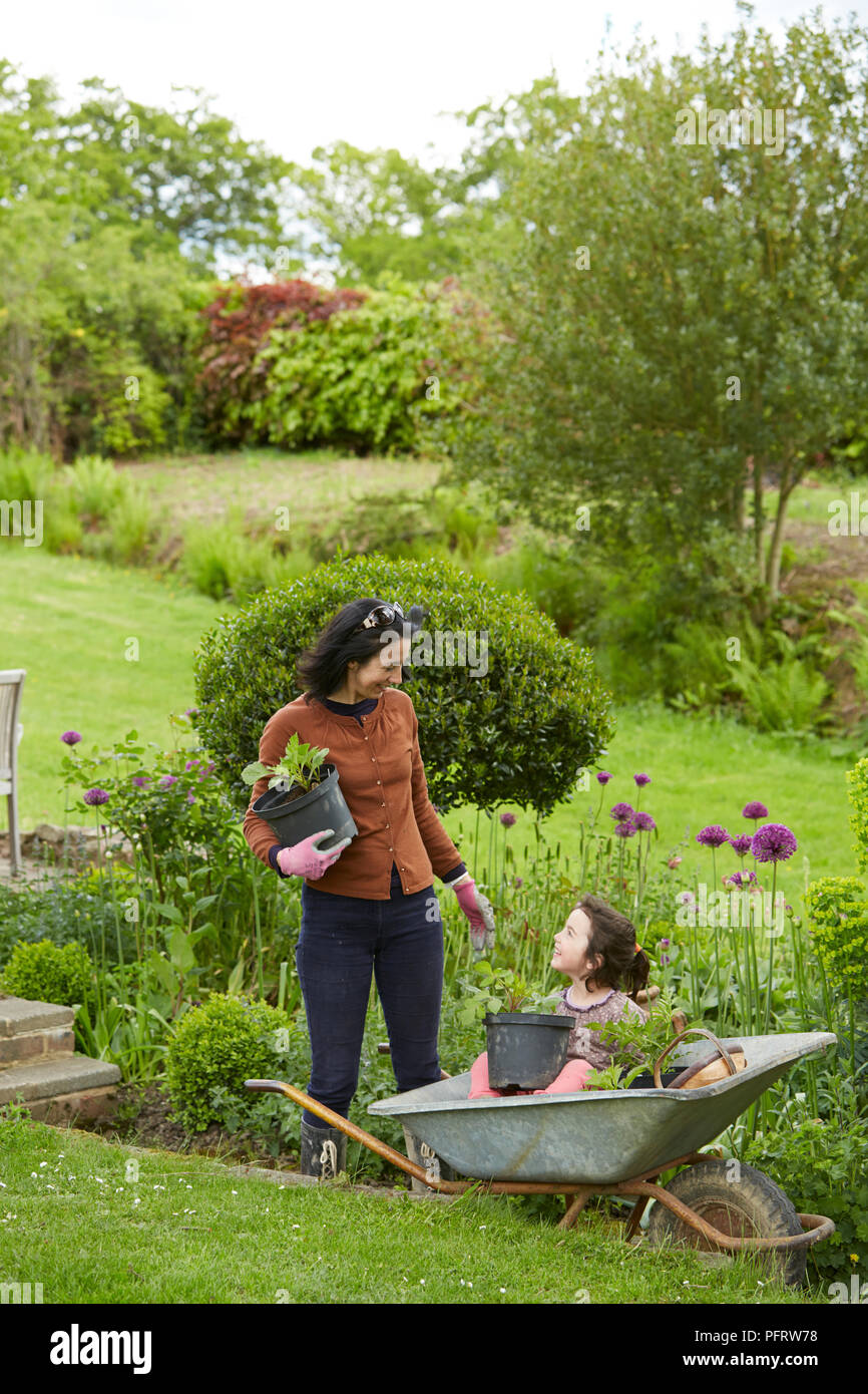 Woman in garden holding plant pot, girl sitting in wheelbarrow next to her Stock Photo
