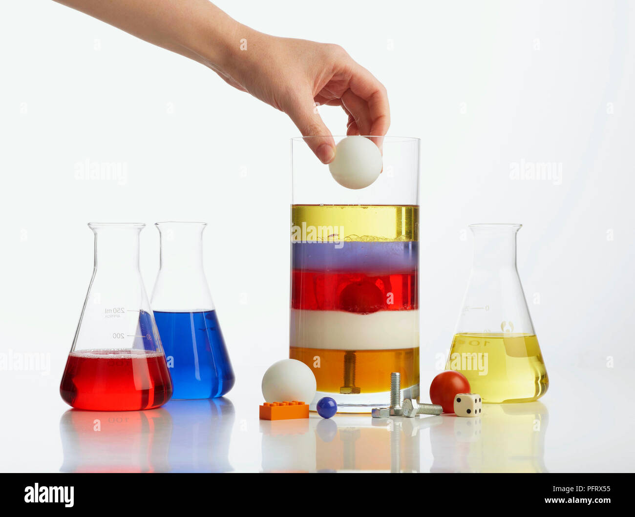 Density tower showing vase with 5 layers: honey, milk, washing-up liquid, water, vegetable oil, and table tennis ball, orange lego block, blue marble, bolts, cherry tomato and dice. Also three large containers with washing-up liquid, water, and vegetable oil. Hand is about to drop table tennis ball into vase Stock Photo